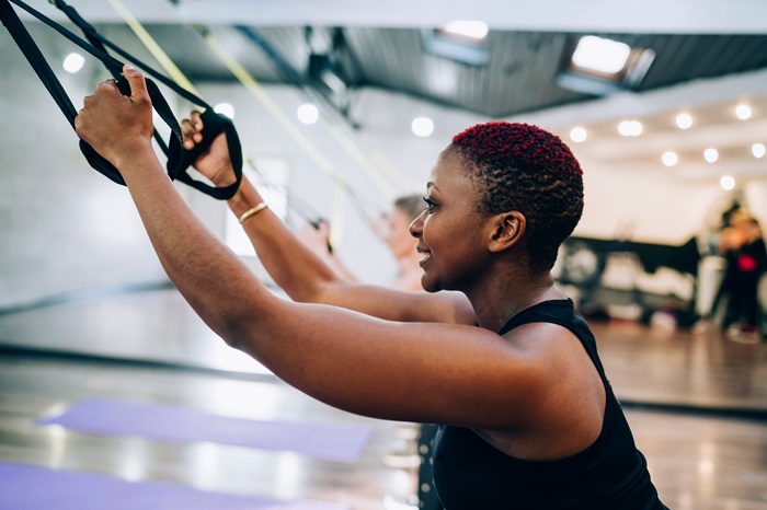 Side view of African American short haired middle aged female in black shirt holding handles of expander in fitness studio
